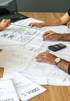 architectural drawings spread on a table with several people's hands, a calculator, and a laptop